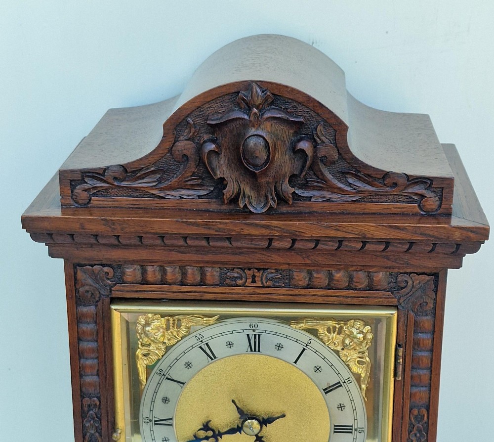 A carved oak mantel clock featuring an arched pediment, ornate brass dial with a silvered chapter ring, and a carved decorative base. View VII