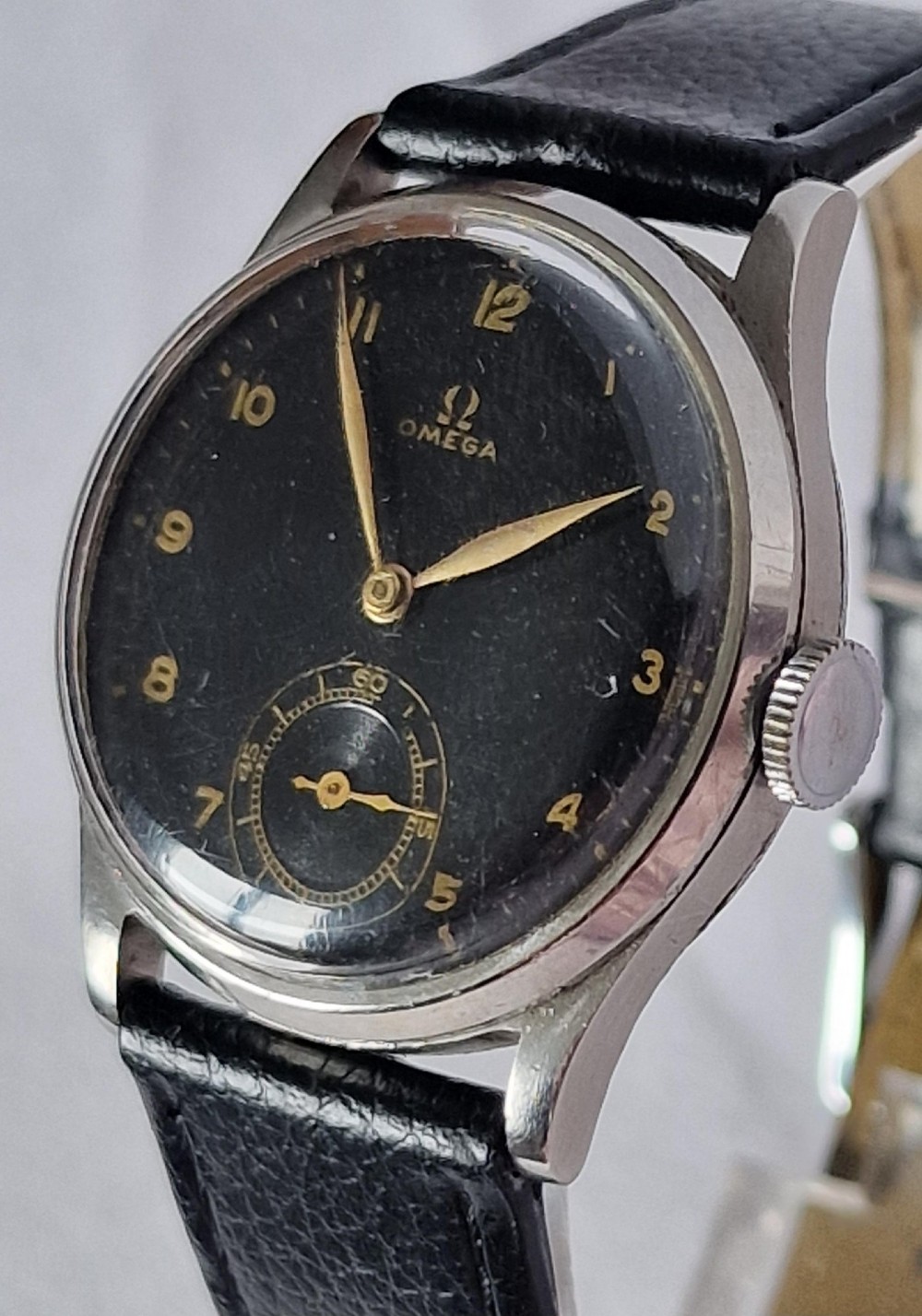 A vintage 1944 Omega wristwatch featuring a circular stainless steel case, black dial with Arabic numerals, sub-seconds dial, and a black leather strap. View VI