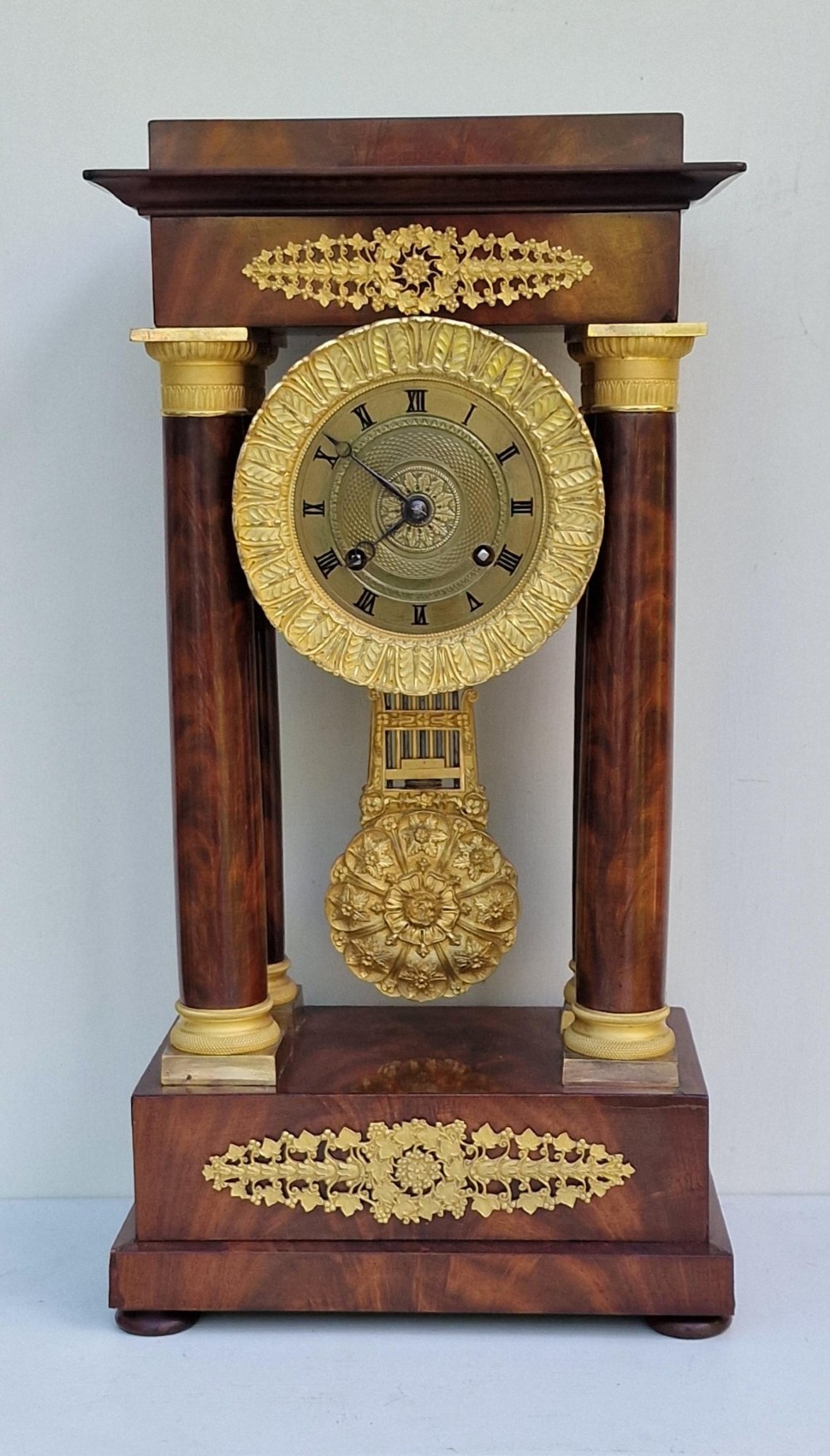 An antique French Empire portico clock featuring four mahogany columns with ornate gilt bronze capitals, an exposed decorative pendulum, and a gilded dial set within a mahogany framework.