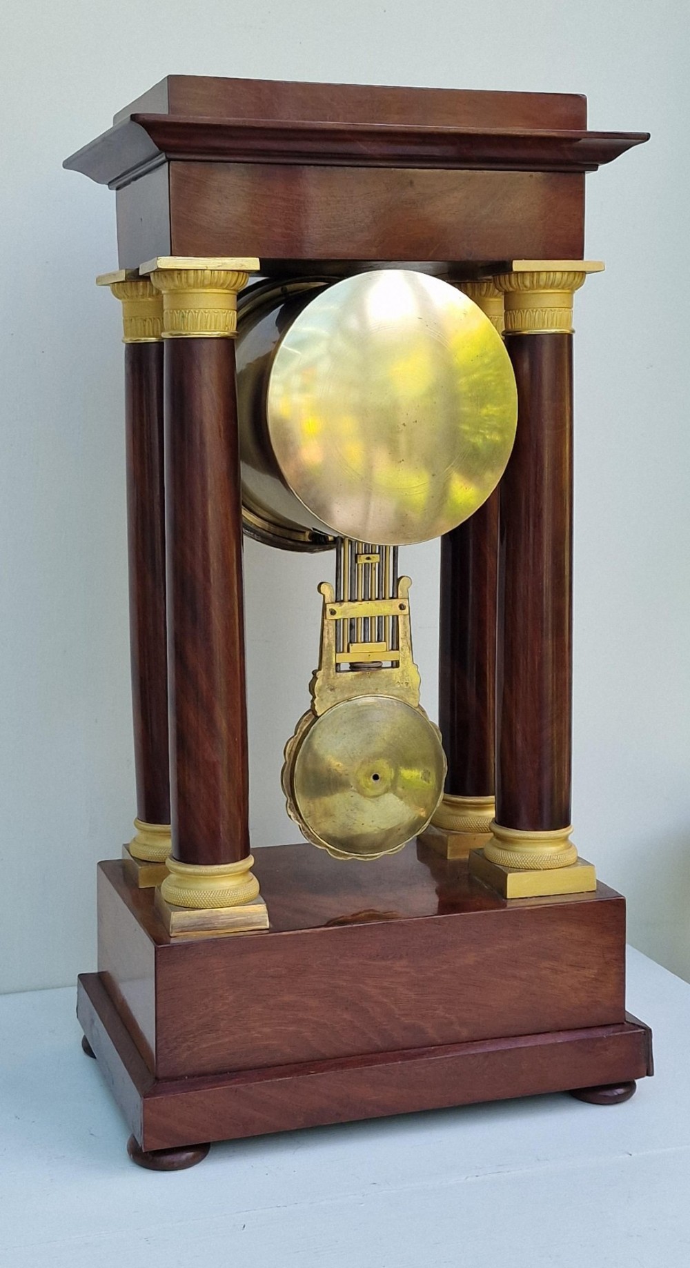 An antique French Empire portico clock featuring four mahogany columns with ornate gilt bronze capitals, an exposed decorative pendulum, and a gilded dial set within a mahogany framework. View II