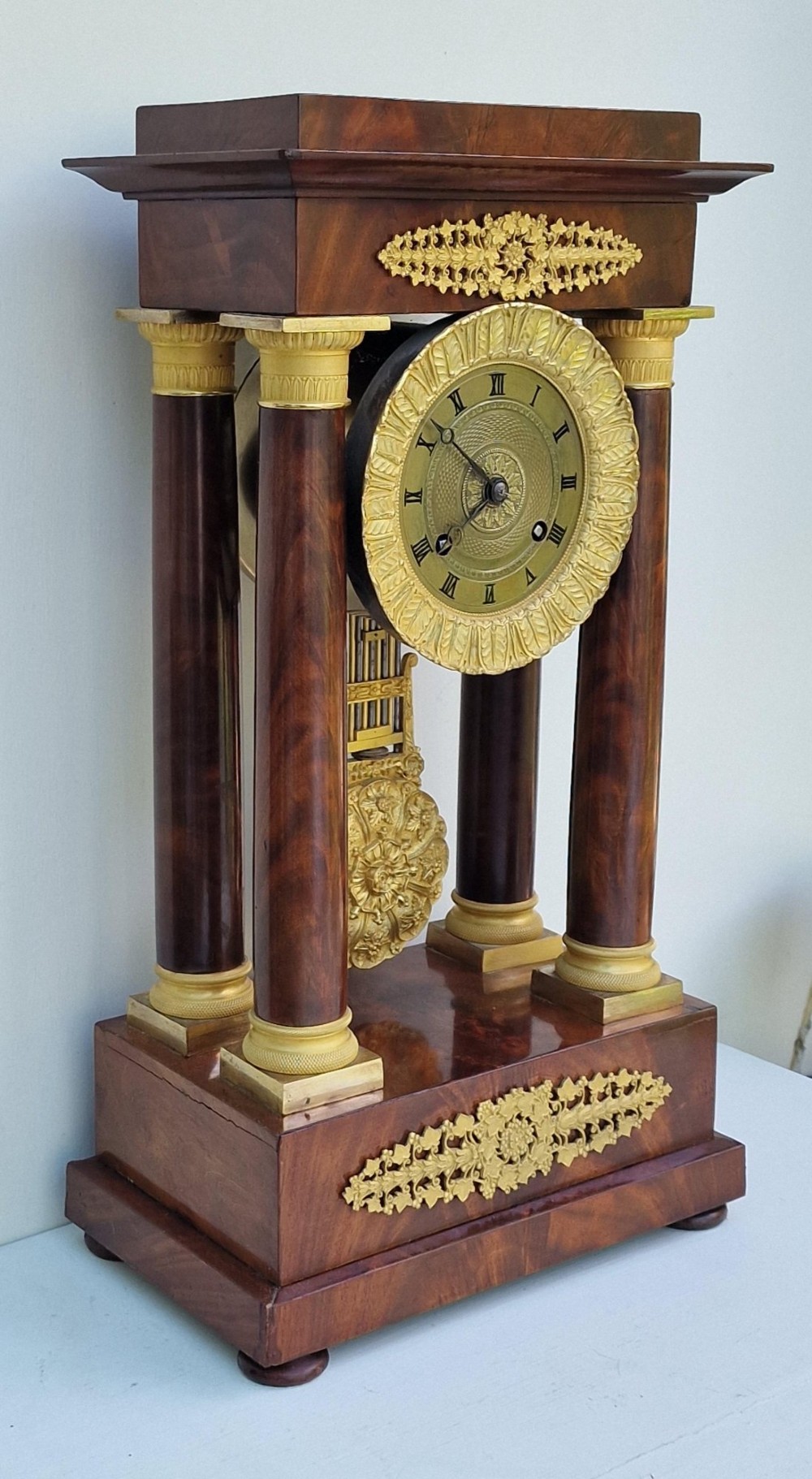 An antique French Empire portico clock featuring four mahogany columns with ornate gilt bronze capitals, an exposed decorative pendulum, and a gilded dial set within a mahogany framework. View IV