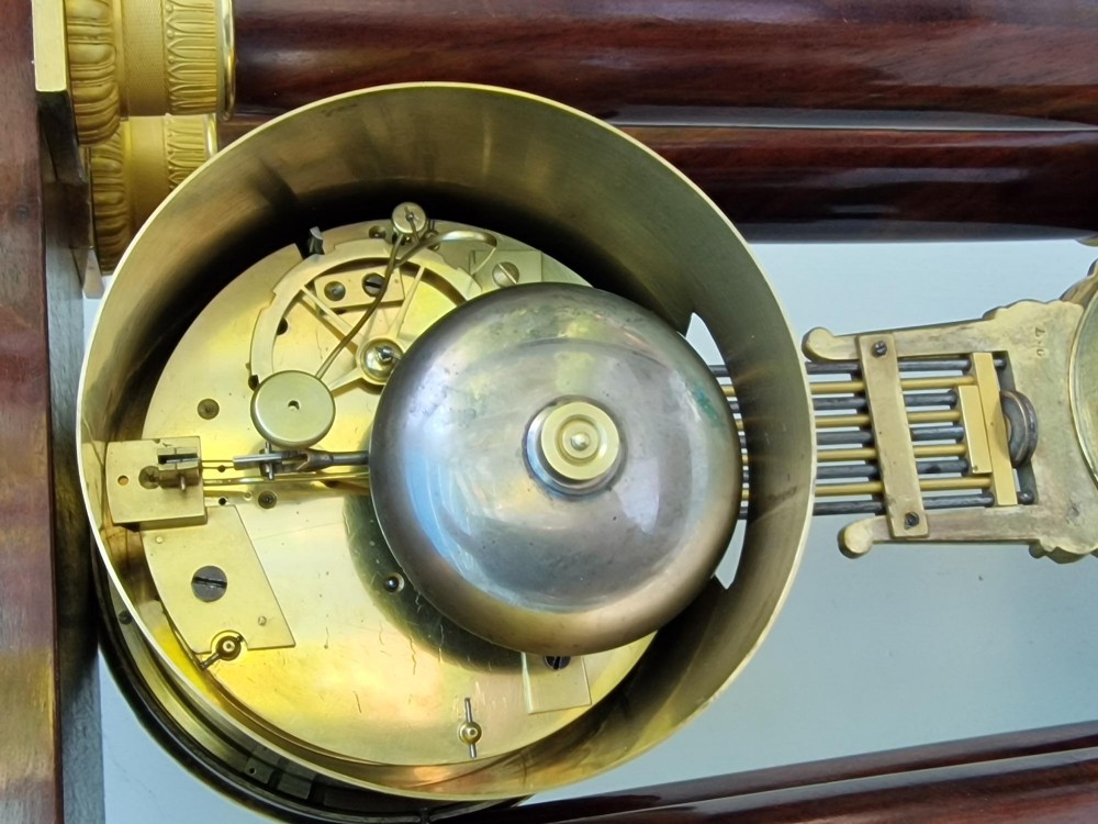 An antique French Empire portico clock featuring four mahogany columns with ornate gilt bronze capitals, an exposed decorative pendulum, and a gilded dial set within a mahogany framework. View V