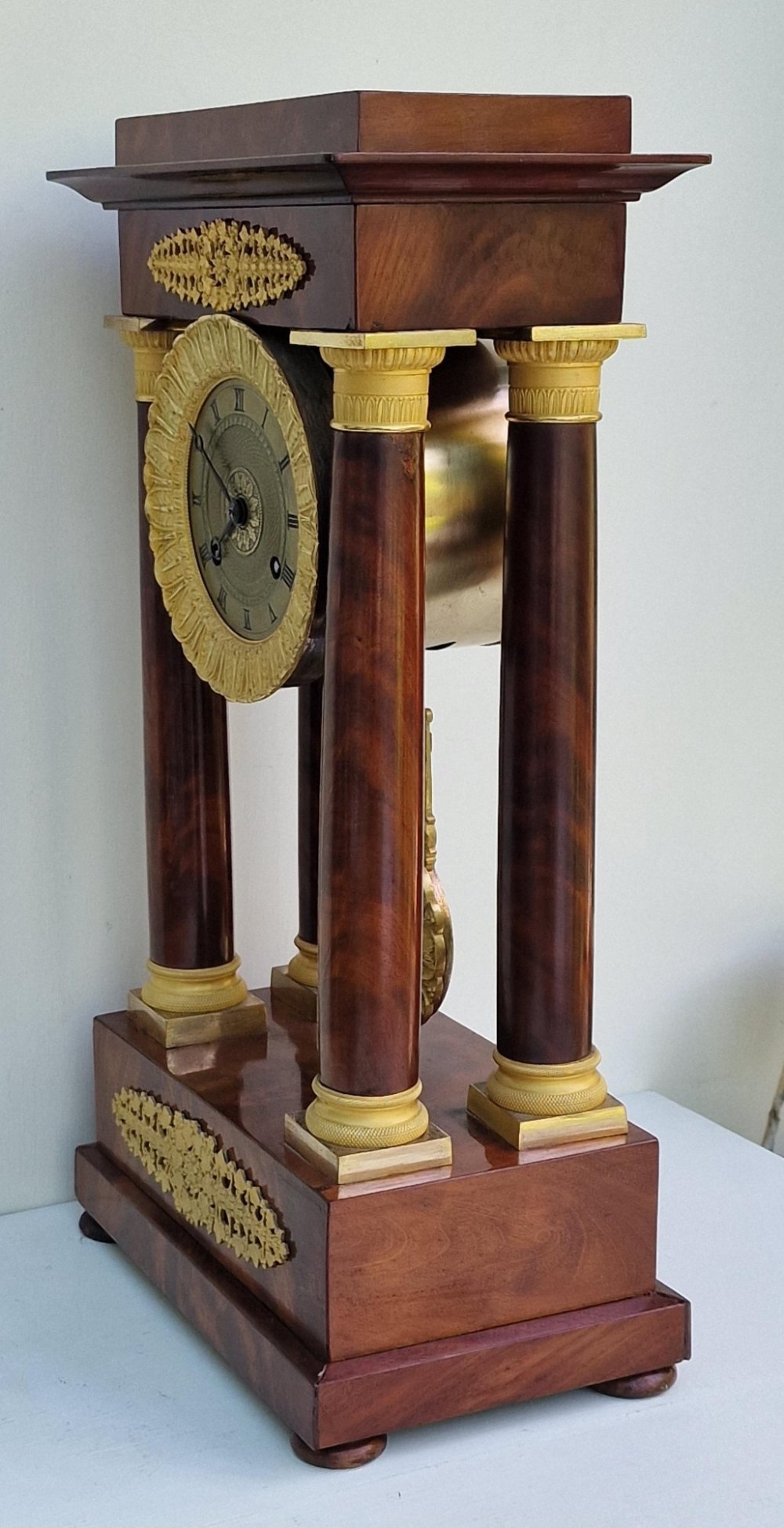 An antique French Empire portico clock featuring four mahogany columns with ornate gilt bronze capitals, an exposed decorative pendulum, and a gilded dial set within a mahogany framework. View VI