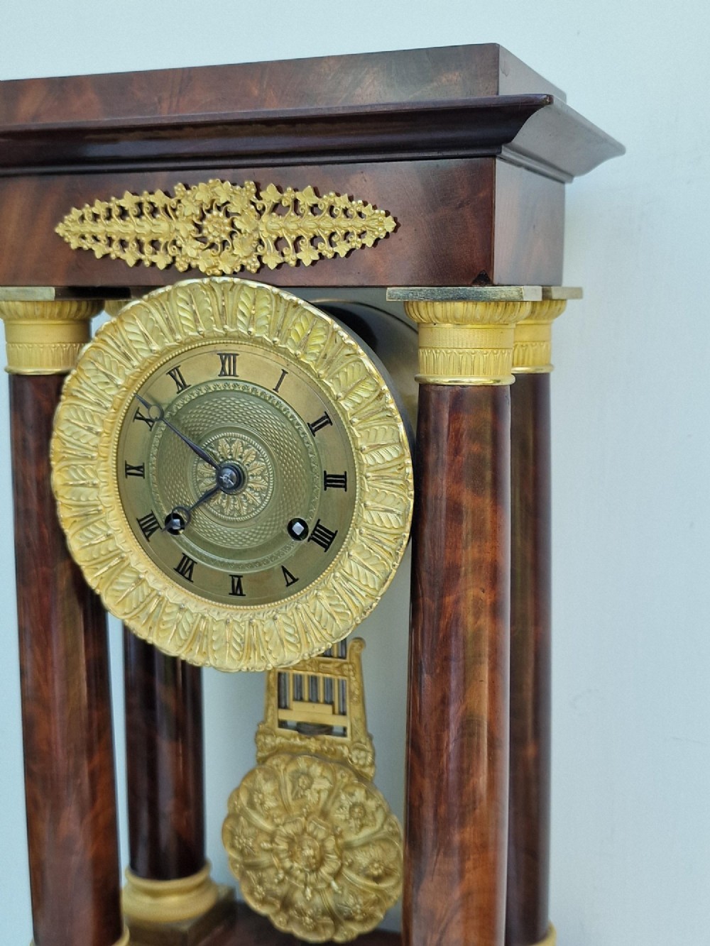 An antique French Empire portico clock featuring four mahogany columns with ornate gilt bronze capitals, an exposed decorative pendulum, and a gilded dial set within a mahogany framework. View VII