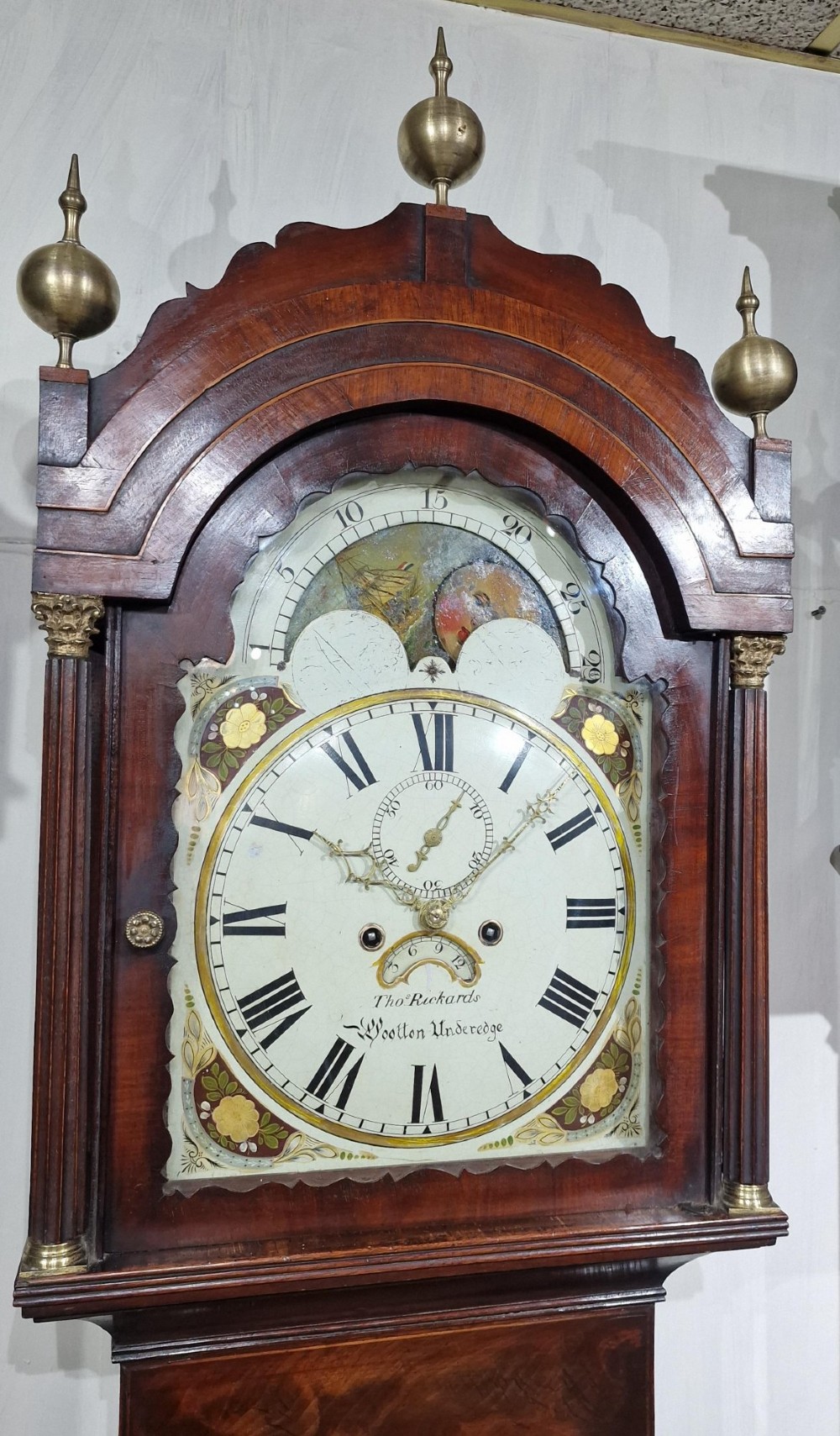 An antique Georgian flame mahogany longcase clock with three brass finials, an arched hood, fluted columns, and a painted dial displaying a moonphase mechanism. View V