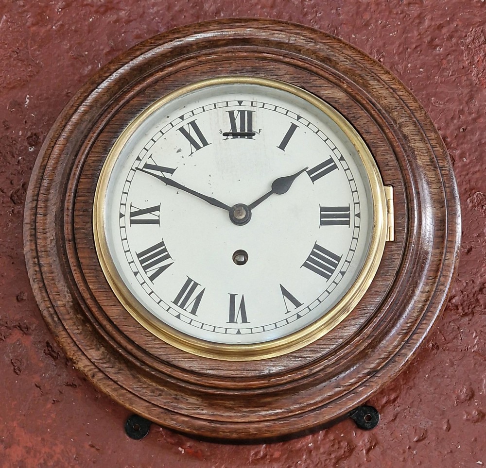 A circular antique oak wall clock featuring a white face with Roman numerals, a brass bezel, and a moulded wood surround.