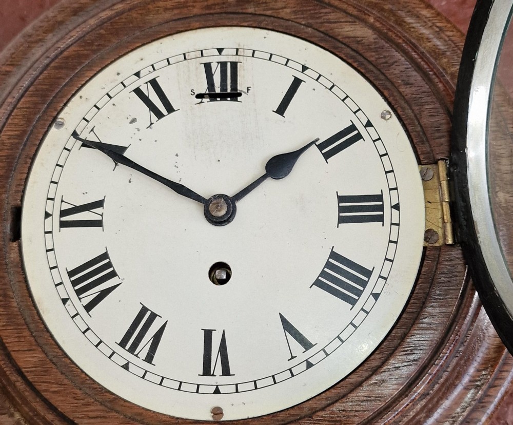 A circular antique oak wall clock featuring a white face with Roman numerals, a brass bezel, and a moulded wood surround. View III