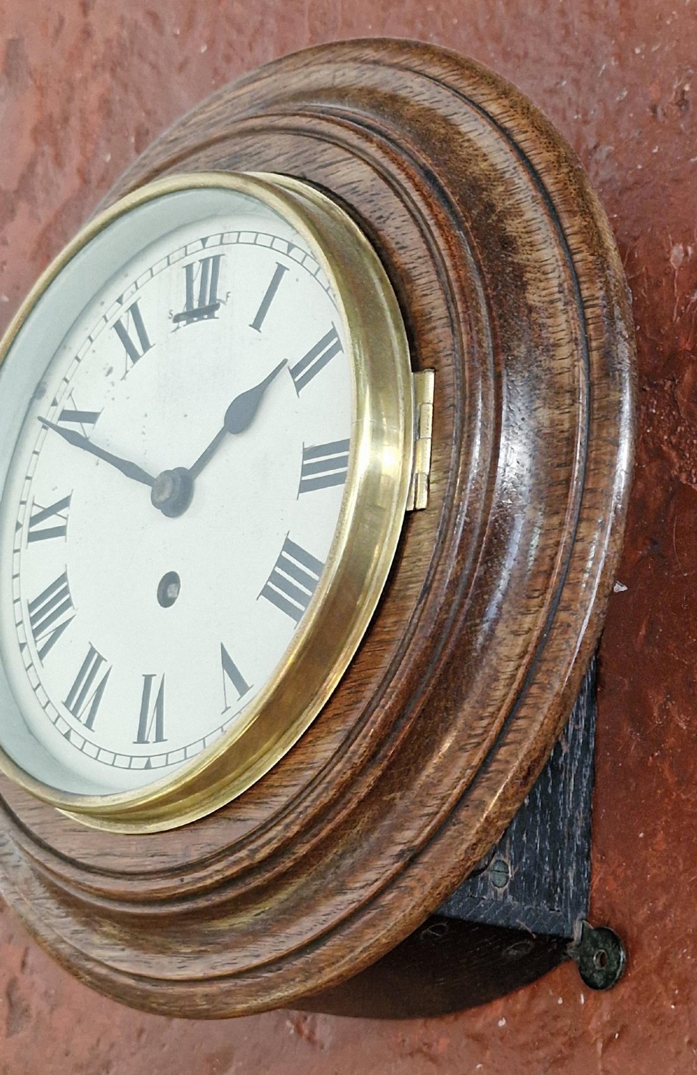 A circular antique oak wall clock featuring a white face with Roman numerals, a brass bezel, and a moulded wood surround. View V