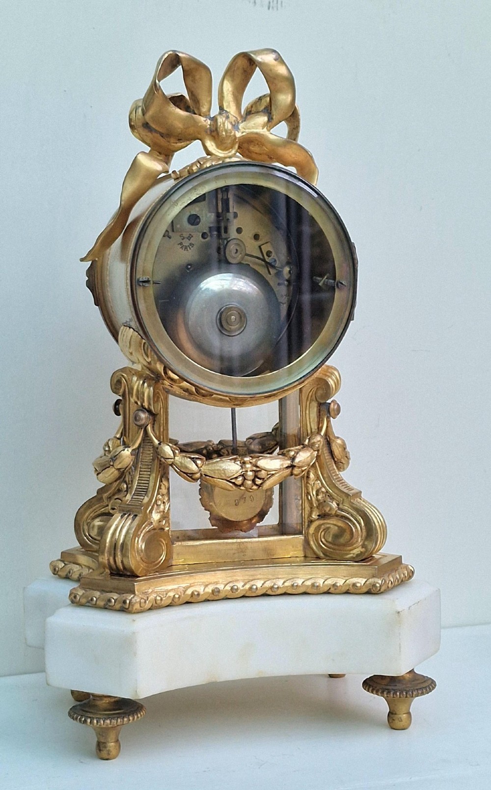 A French gilt bronze mantel clock featuring a white enamel dial with Arabic numerals, an ornate ribbon bow crest, floral swags, and a white marble plinth base. View II