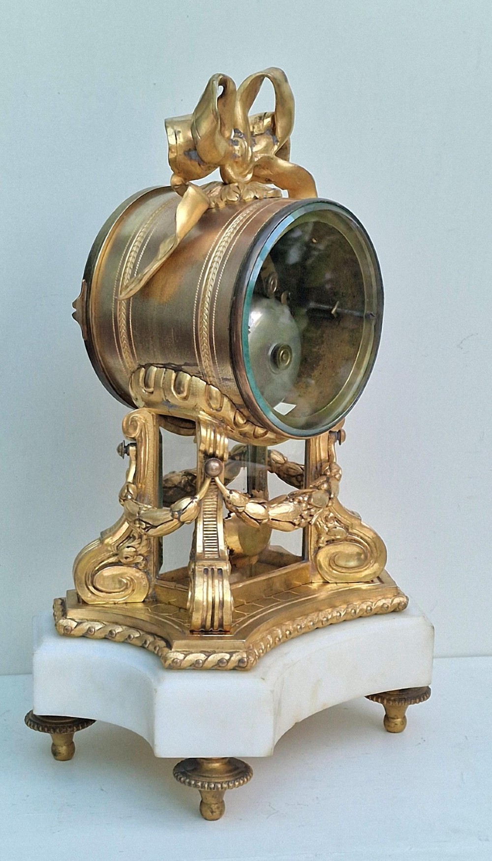 A French gilt bronze mantel clock featuring a white enamel dial with Arabic numerals, an ornate ribbon bow crest, floral swags, and a white marble plinth base. View III