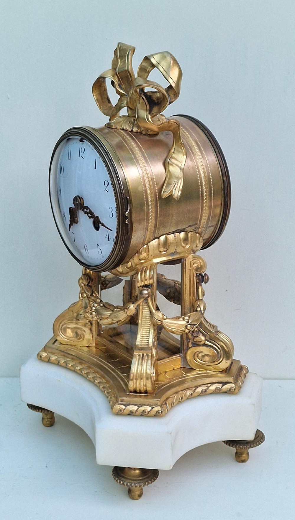 A French gilt bronze mantel clock featuring a white enamel dial with Arabic numerals, an ornate ribbon bow crest, floral swags, and a white marble plinth base. View VIII
