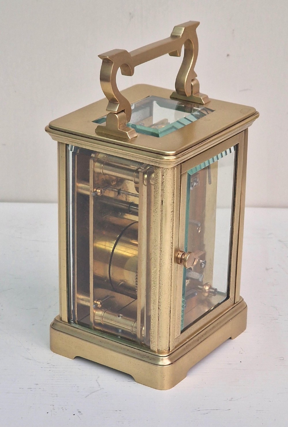 A rectangular brass French carriage clock featuring bevelled glass panels, a white enamel dial with black Roman numerals, and a top carrying handle. View V