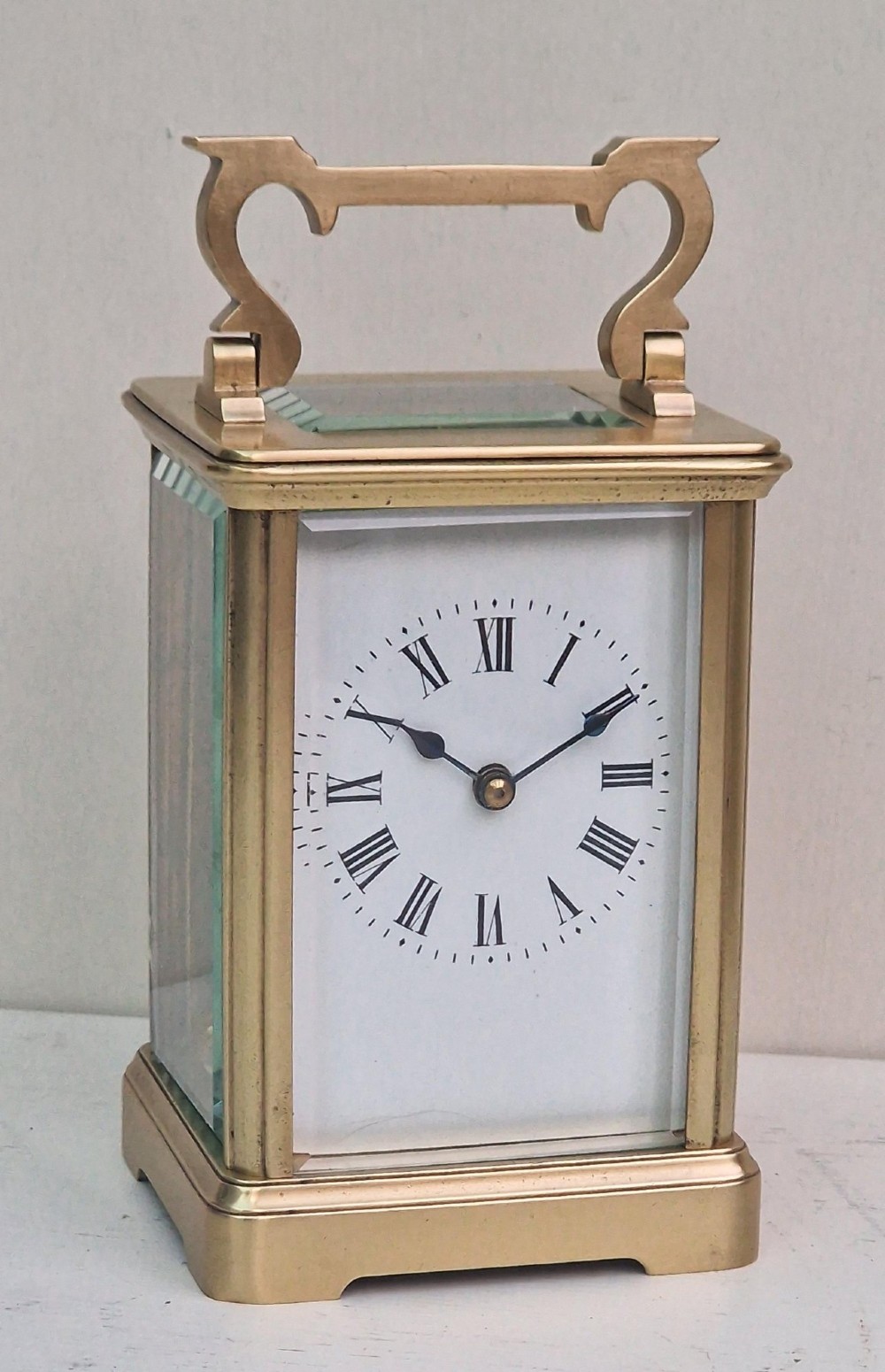 A rectangular brass French carriage clock featuring bevelled glass panels, a white enamel dial with black Roman numerals, and a top carrying handle. View VI