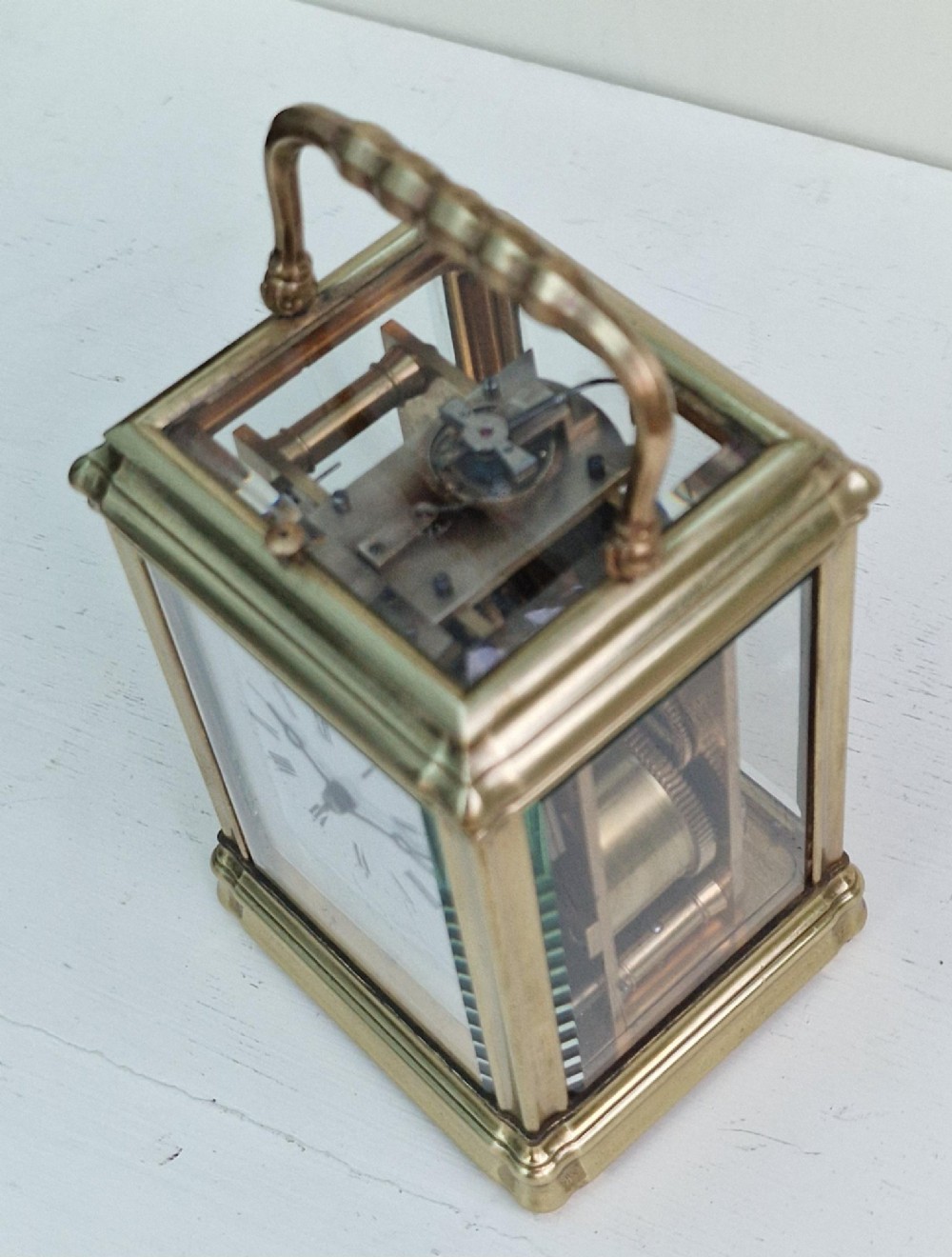 An antique French brass carriage clock featuring a gorge case structure, bevelled glass side panels, a white enamel dial with Roman numerals, and a top carrying handle. View VII