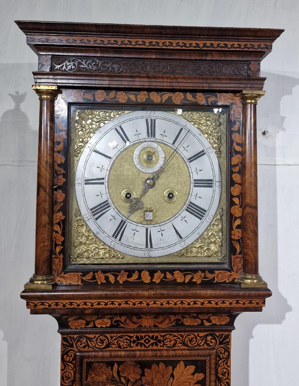 An English late 17th-century longcase clock with intricate floral marquetry veneer, a square brass dial, and a circular glass lenticle on the trunk door. View V