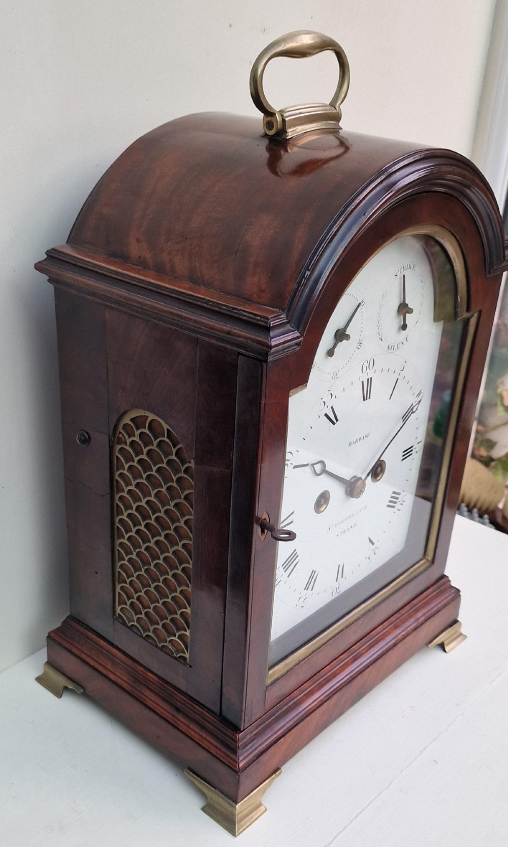 An antique Georgian mahogany bracket clock featuring an arched top with a brass carrying handle, brass bracket feet, and a white dial inscribed Barwise London. View V