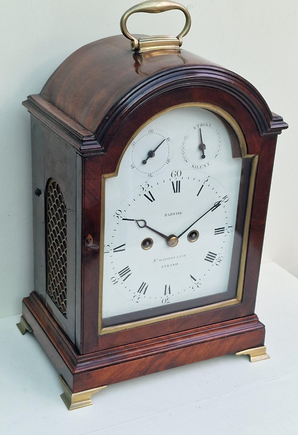 An antique Georgian mahogany bracket clock featuring an arched top with a brass carrying handle, brass bracket feet, and a white dial inscribed Barwise London. View VIII