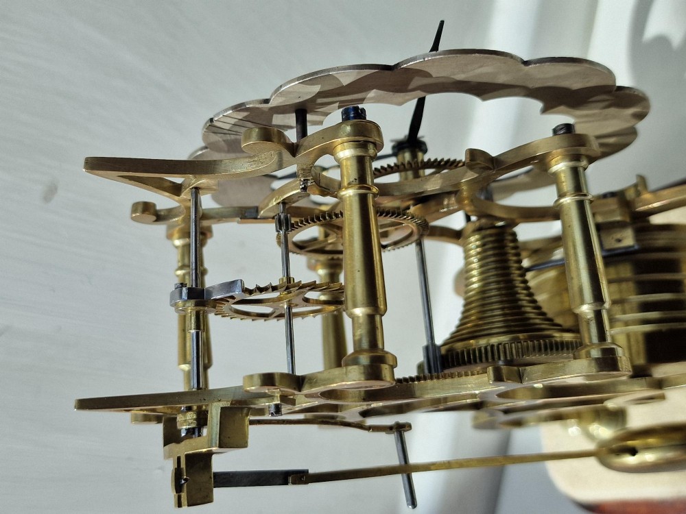 A Victorian brass skeleton clock displaying exposed gearwork and a silvered chapter ring with Roman numerals, set on a plinth under a clear glass dome. View VI