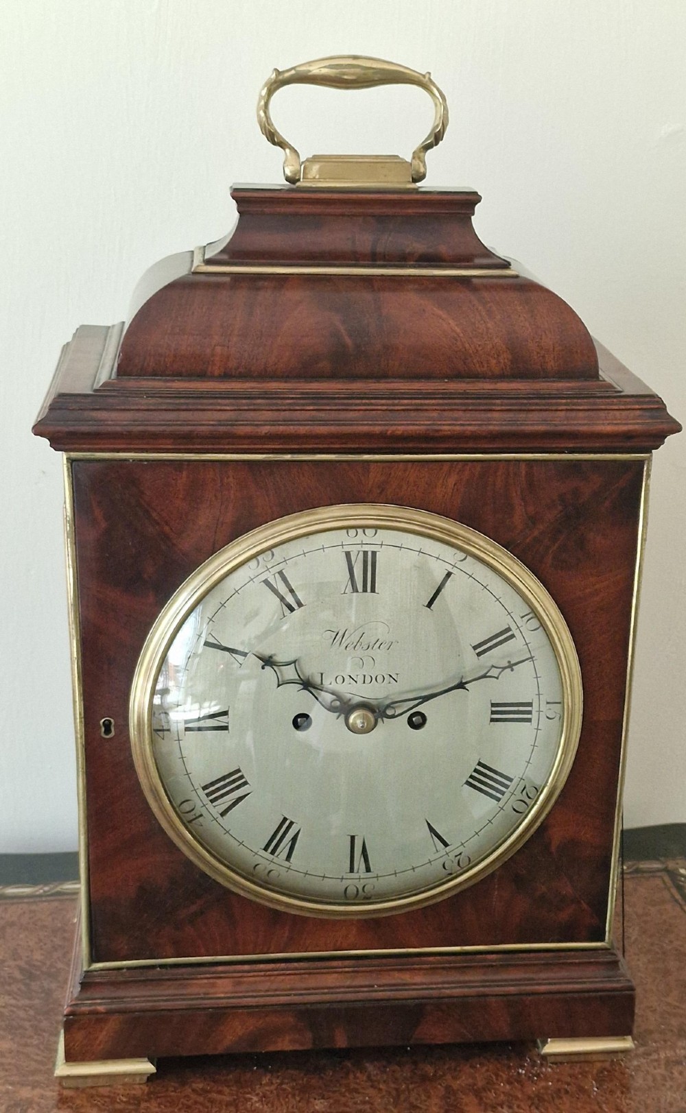 A Georgian flame mahogany bracket clock featuring a brass carrying handle, circular white dial with Roman numerals, and brass-backed side frets. View II