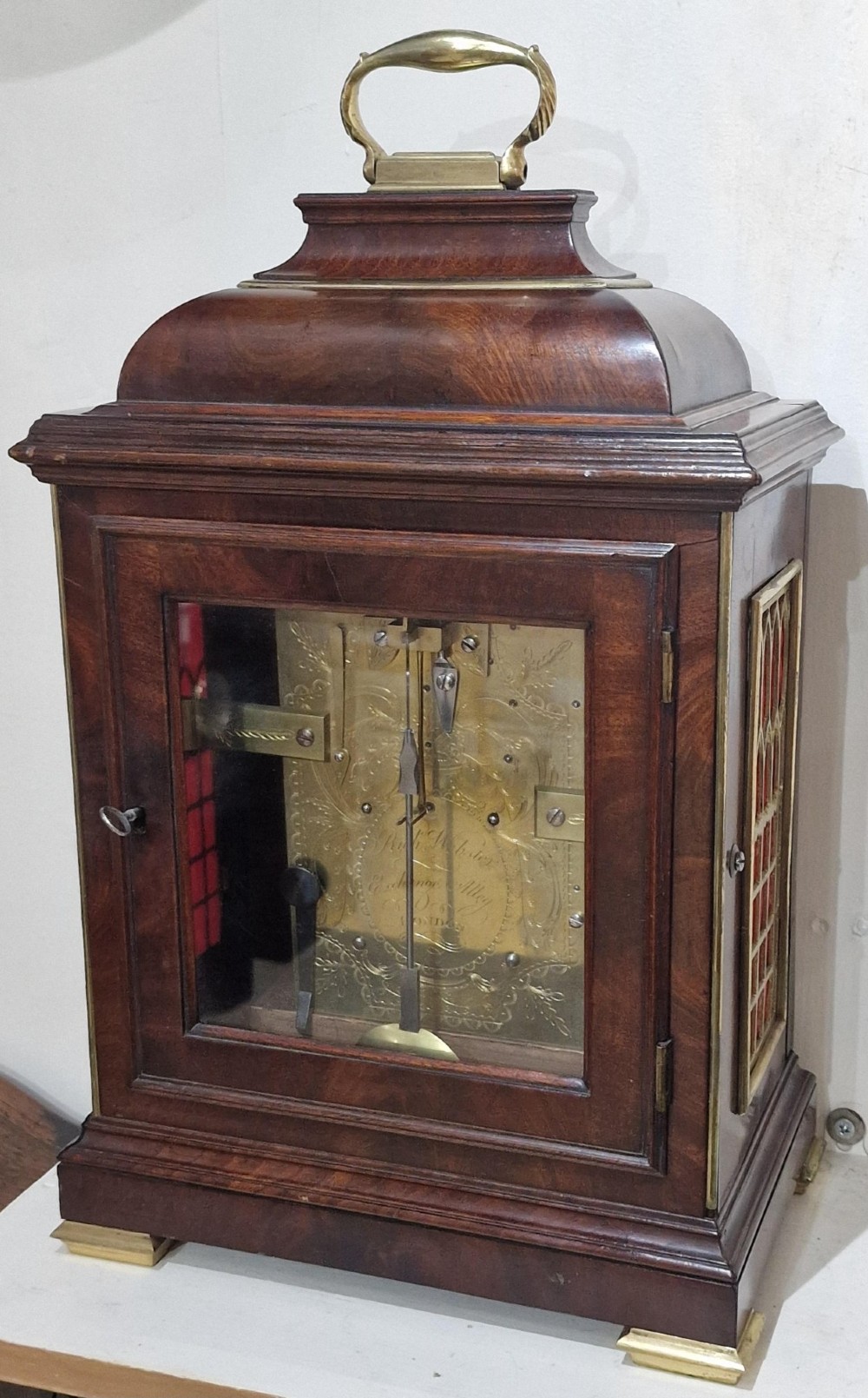 A Georgian flame mahogany bracket clock featuring a brass carrying handle, circular white dial with Roman numerals, and brass-backed side frets. View III
