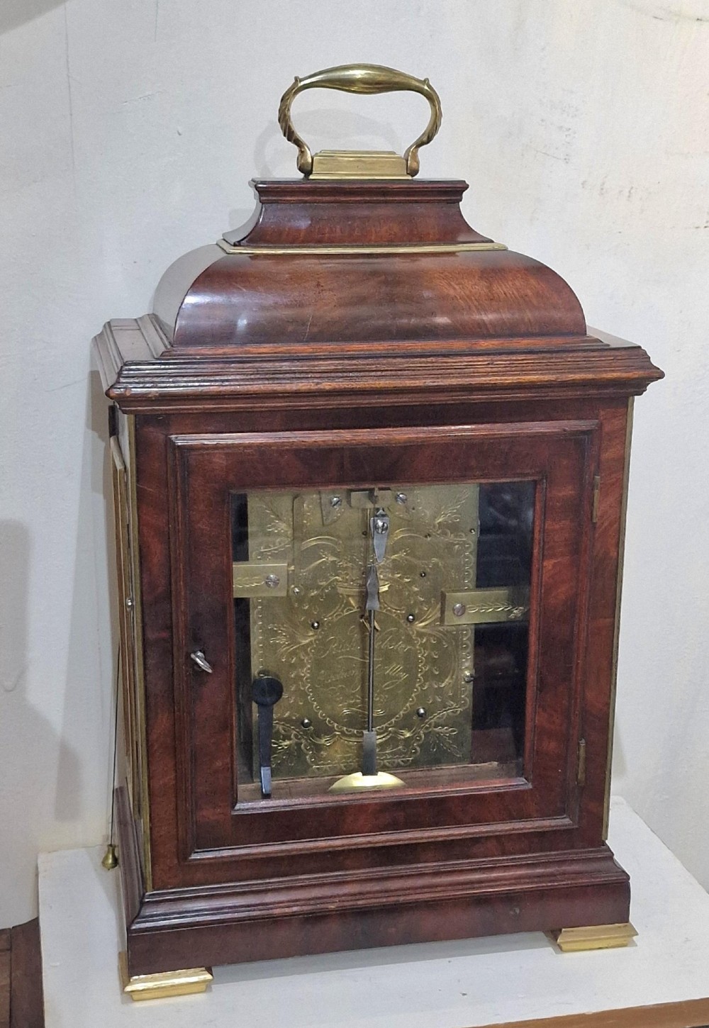 A Georgian flame mahogany bracket clock featuring a brass carrying handle, circular white dial with Roman numerals, and brass-backed side frets. View IV