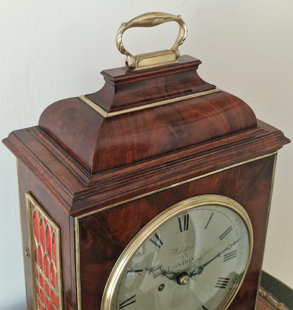 A Georgian flame mahogany bracket clock featuring a brass carrying handle, circular white dial with Roman numerals, and brass-backed side frets. View X