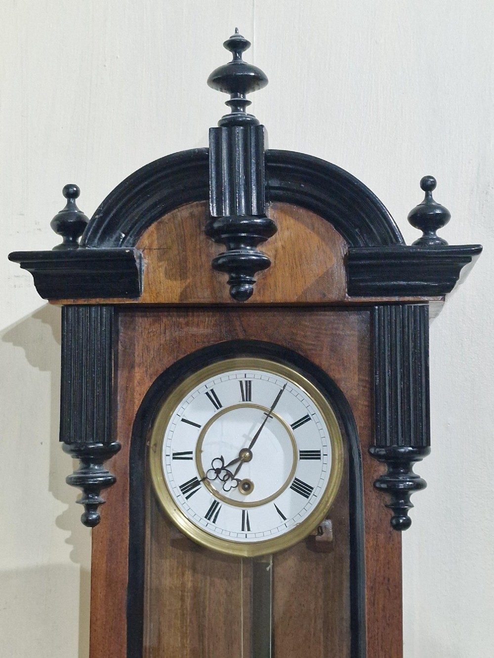 An Austrian Vienna regulator wall clock featuring a glazed walnut case with ebonised architectural columns, turned finials, a white Roman numeral dial, and a brass pendulum. View VI