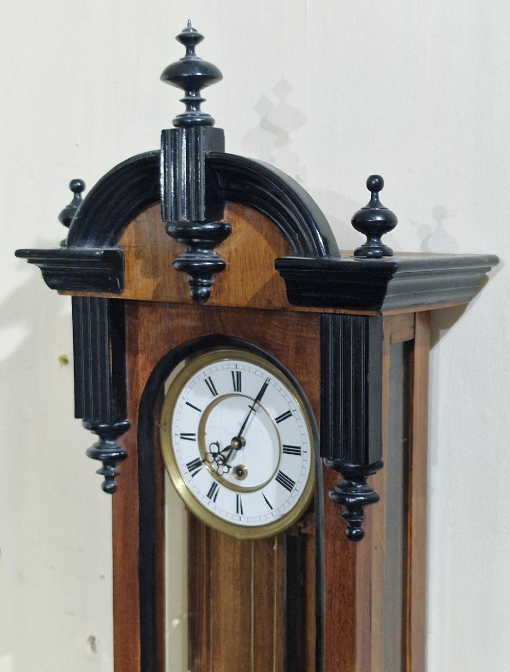 An Austrian Vienna regulator wall clock featuring a glazed walnut case with ebonised architectural columns, turned finials, a white Roman numeral dial, and a brass pendulum. View VII