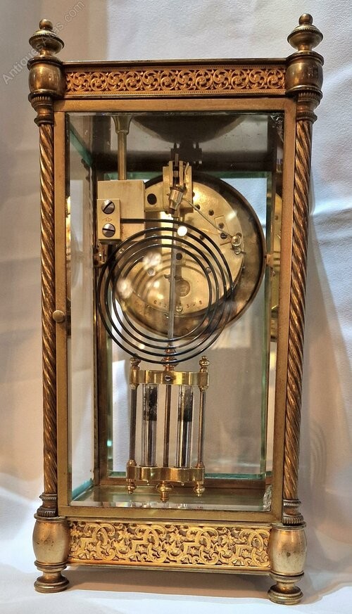 A brass four-glass mantel clock featuring twisted corner columns, decorative filigree friezes, a circular dial decorated with a cherub, and a visible twin mercury pendulum. View II
