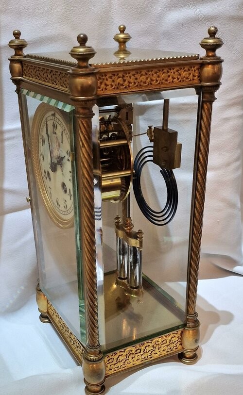 A brass four-glass mantel clock featuring twisted corner columns, decorative filigree friezes, a circular dial decorated with a cherub, and a visible twin mercury pendulum. View V