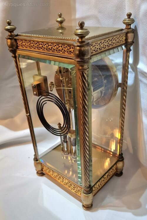 A brass four-glass mantel clock featuring twisted corner columns, decorative filigree friezes, a circular dial decorated with a cherub, and a visible twin mercury pendulum. View VI