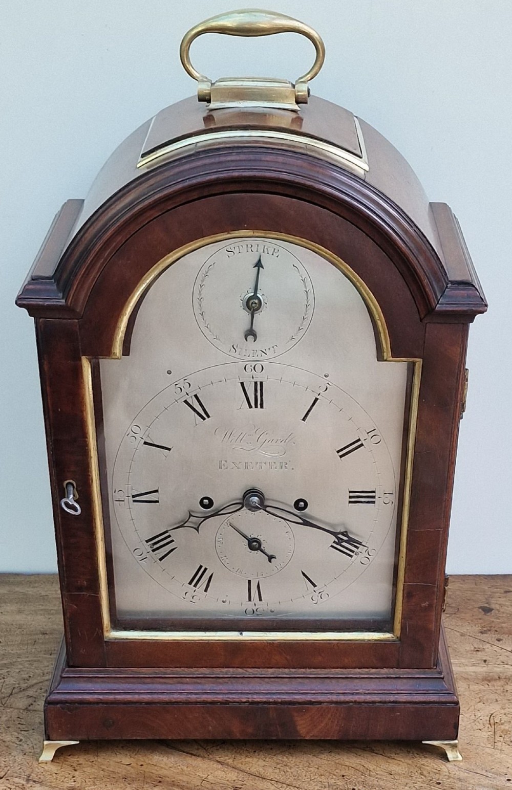 A Georgian mahogany bracket clock featuring an arched top with a brass carrying handle, silvered engraved dial with Roman numerals, and brass bracket feet.