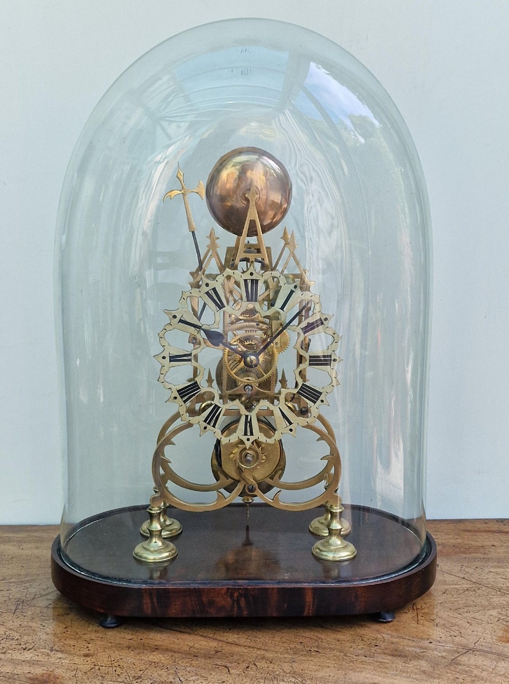 A Victorian brass skeleton clock with an exposed mechanical movement and top strike bell, housed under a clear glass dome on an oval mahogany base. View II