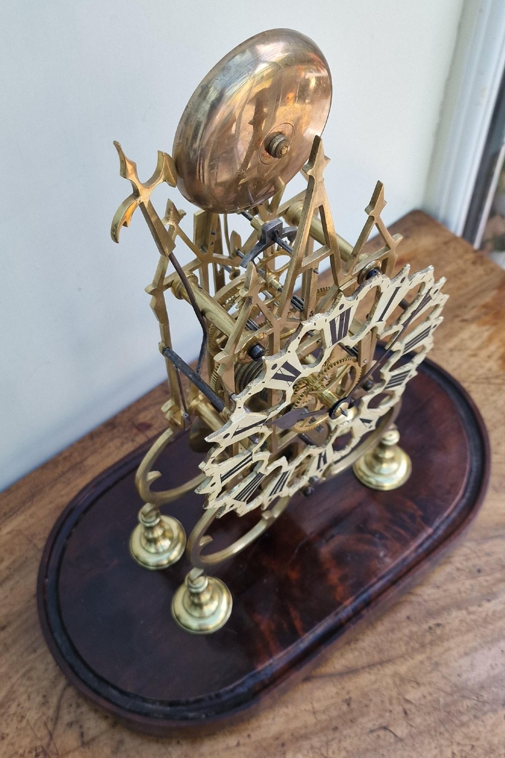 A Victorian brass skeleton clock with an exposed mechanical movement and top strike bell, housed under a clear glass dome on an oval mahogany base. View VI
