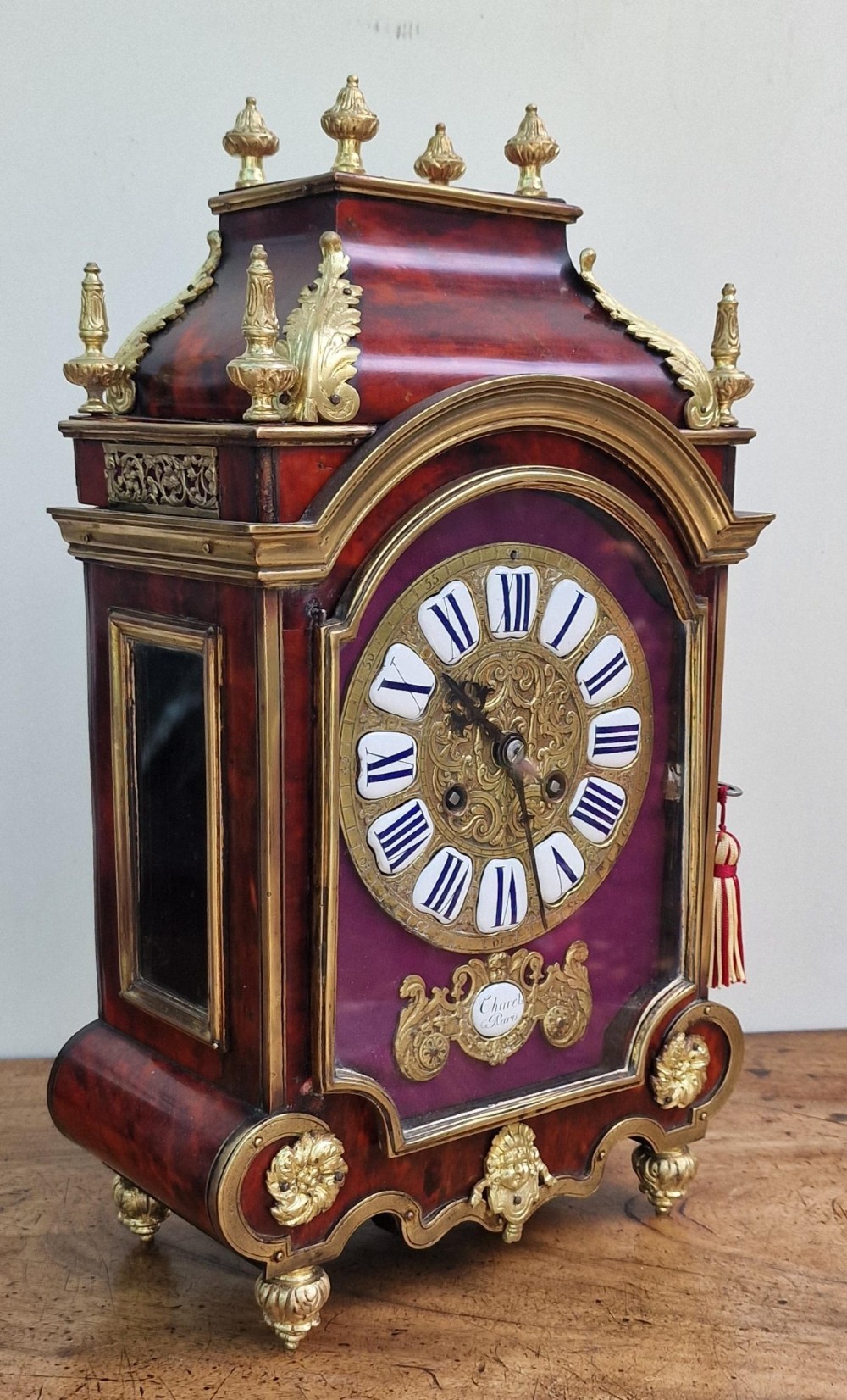 A red tortoiseshell and gilt bronze mantel clock featuring enamel Roman numeral cartouches and cast finials. View IX