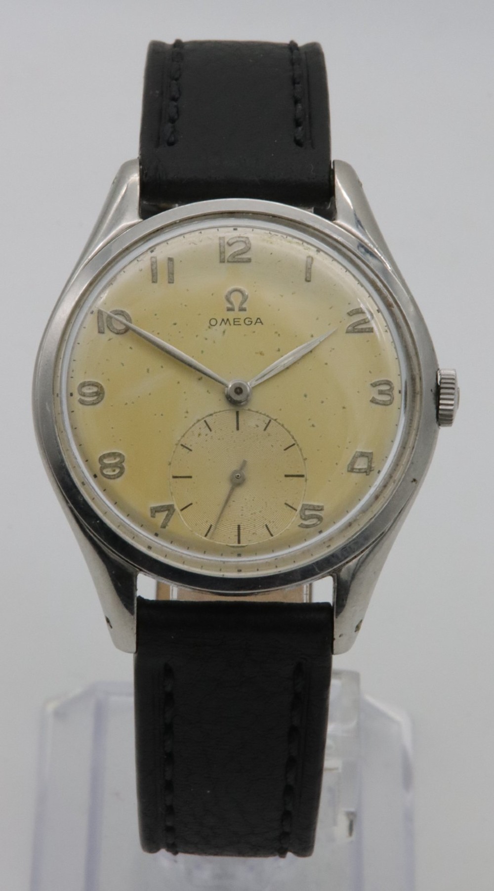 A 1947 Omega wristwatch featuring a circular stainless steel case, a patinated cream dial with Arabic numerals, a sub-seconds dial, and a black leather strap.
