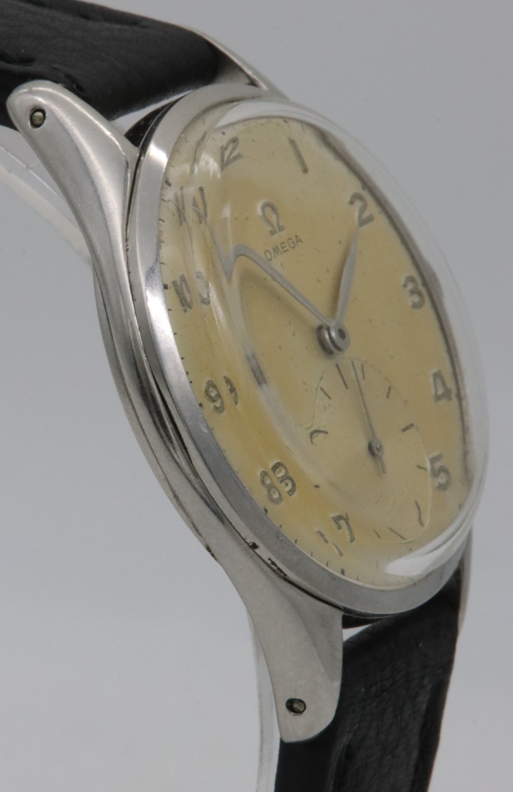 A 1947 Omega wristwatch featuring a circular stainless steel case, a patinated cream dial with Arabic numerals, a sub-seconds dial, and a black leather strap. View V