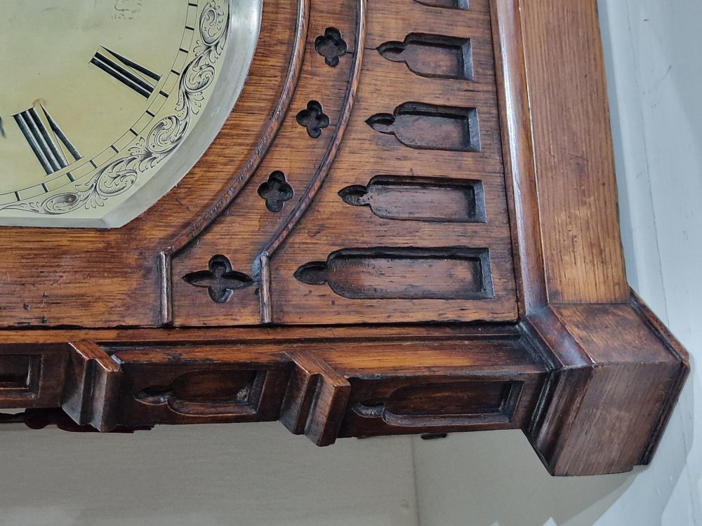 A carved oak Gothic Revival bracket clock featuring pointed arch cresting, turned finials, blind tracery details, and an engraved brass dial with Roman numerals. View VI