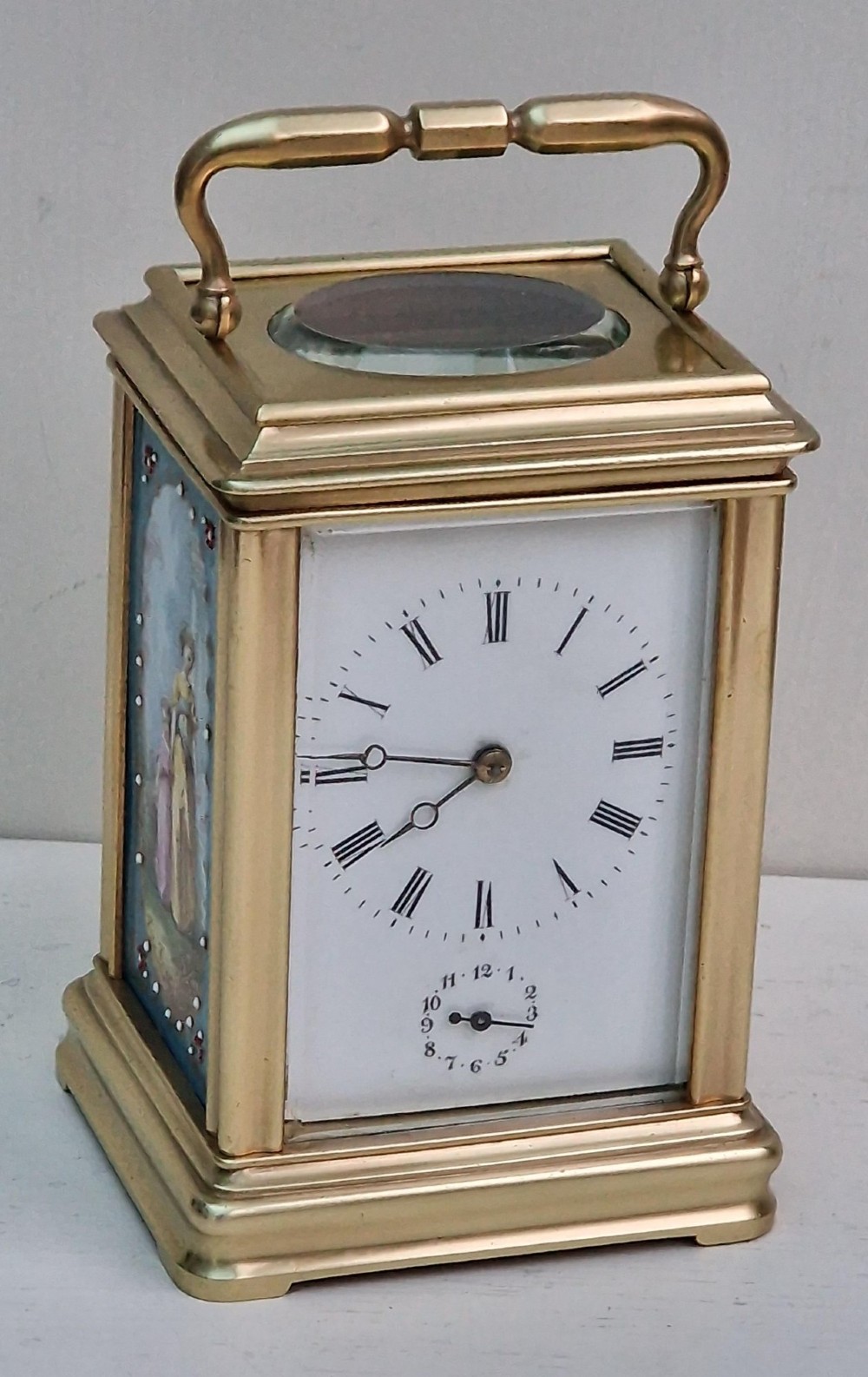 A brass carriage clock featuring a hinged top handle, a glass viewing window above, a white enamel dial with Roman numerals, and side panels decorated with painted porcelain figurative scenes.
