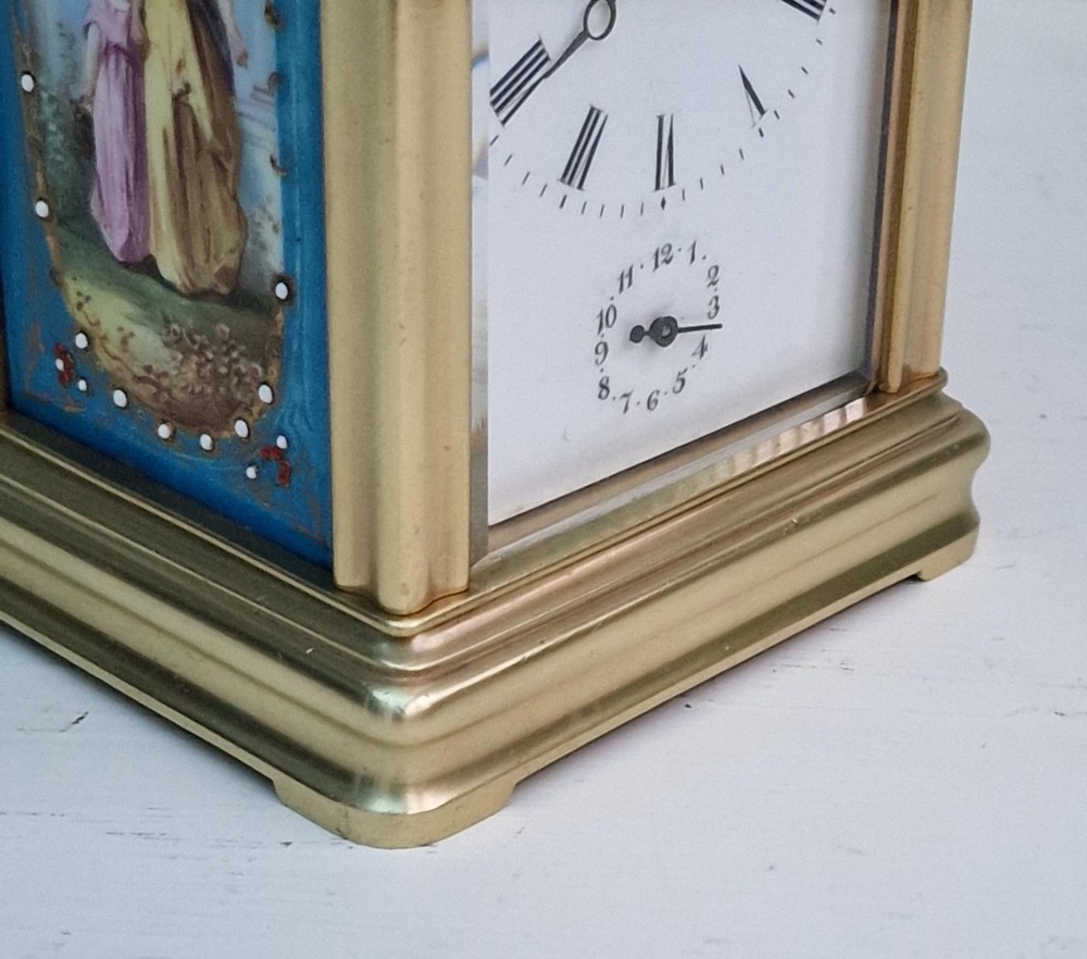 A brass carriage clock featuring a hinged top handle, a glass viewing window above, a white enamel dial with Roman numerals, and side panels decorated with painted porcelain figurative scenes. View II