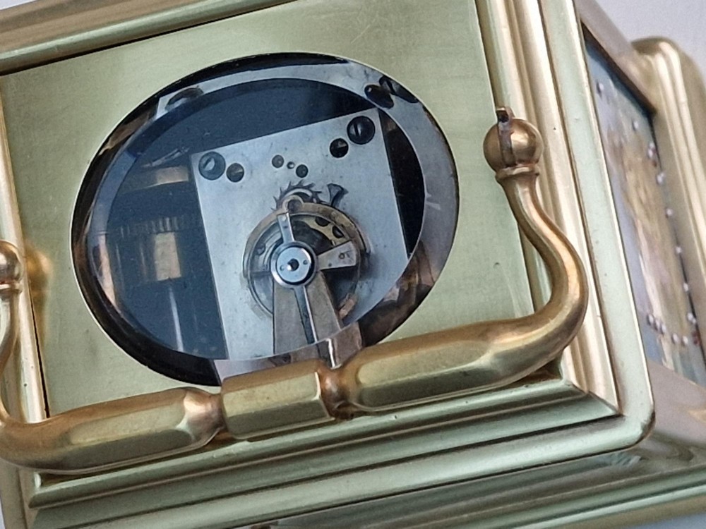 A brass carriage clock featuring a hinged top handle, a glass viewing window above, a white enamel dial with Roman numerals, and side panels decorated with painted porcelain figurative scenes. View III