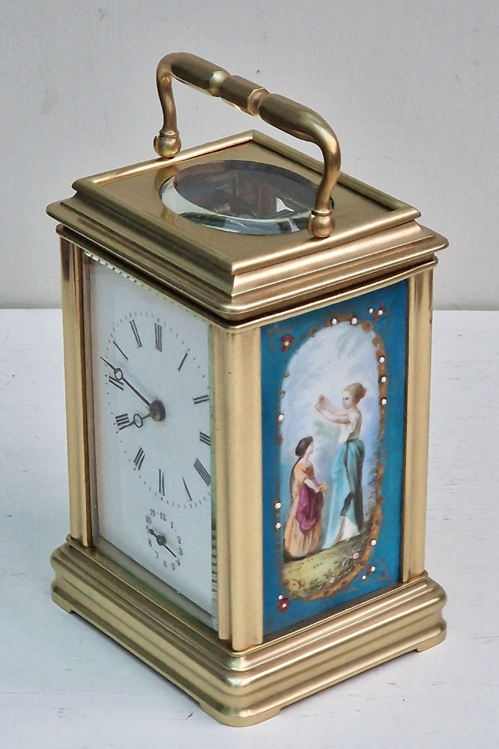 A brass carriage clock featuring a hinged top handle, a glass viewing window above, a white enamel dial with Roman numerals, and side panels decorated with painted porcelain figurative scenes. View IV