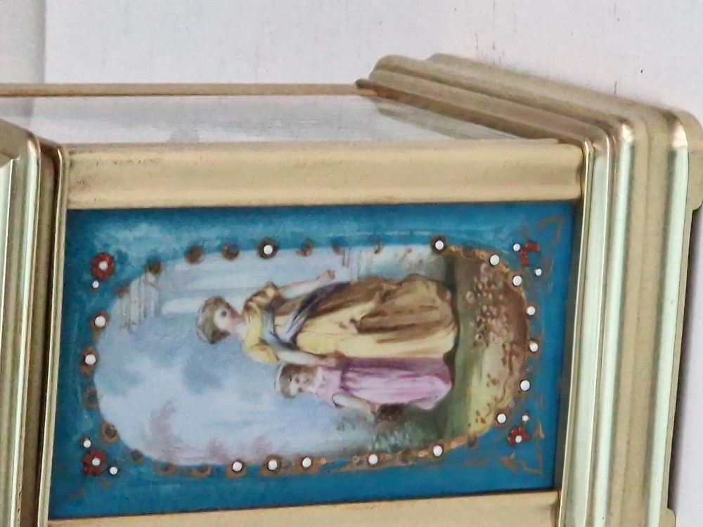A brass carriage clock featuring a hinged top handle, a glass viewing window above, a white enamel dial with Roman numerals, and side panels decorated with painted porcelain figurative scenes. View VI