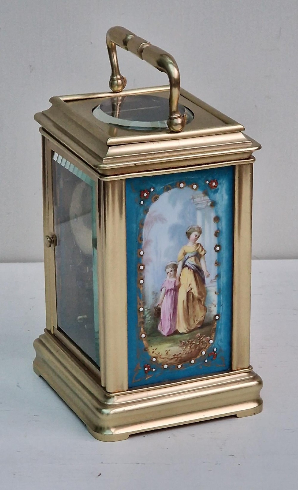 A brass carriage clock featuring a hinged top handle, a glass viewing window above, a white enamel dial with Roman numerals, and side panels decorated with painted porcelain figurative scenes. View VII