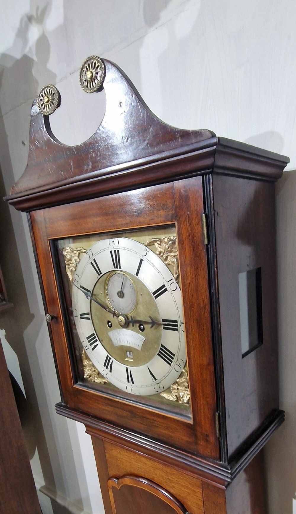 A Georgian mahogany grandmother clock featuring a swan-neck pediment, brass rosettes, an engraved brass and silvered dial, and an inlaid wooden trunk case. View IV