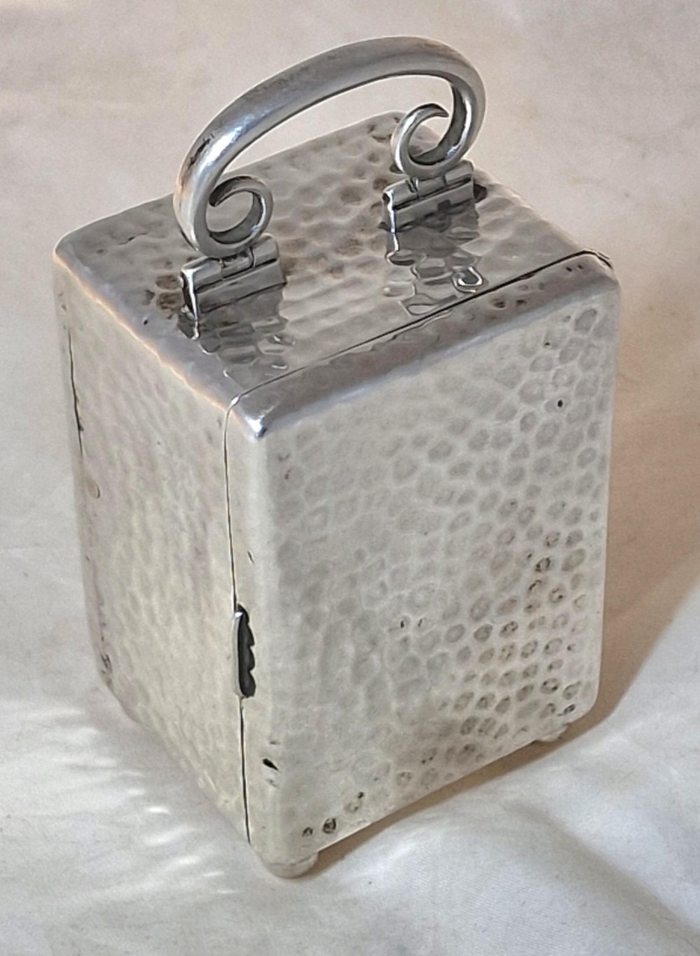 An Edwardian sterling silver carriage clock featuring a planished hammered case, scrolling carrying handle, ball feet, and a white dial with Roman numerals. View II