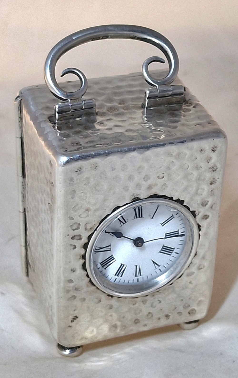 An Edwardian sterling silver carriage clock featuring a planished hammered case, scrolling carrying handle, ball feet, and a white dial with Roman numerals. View IV