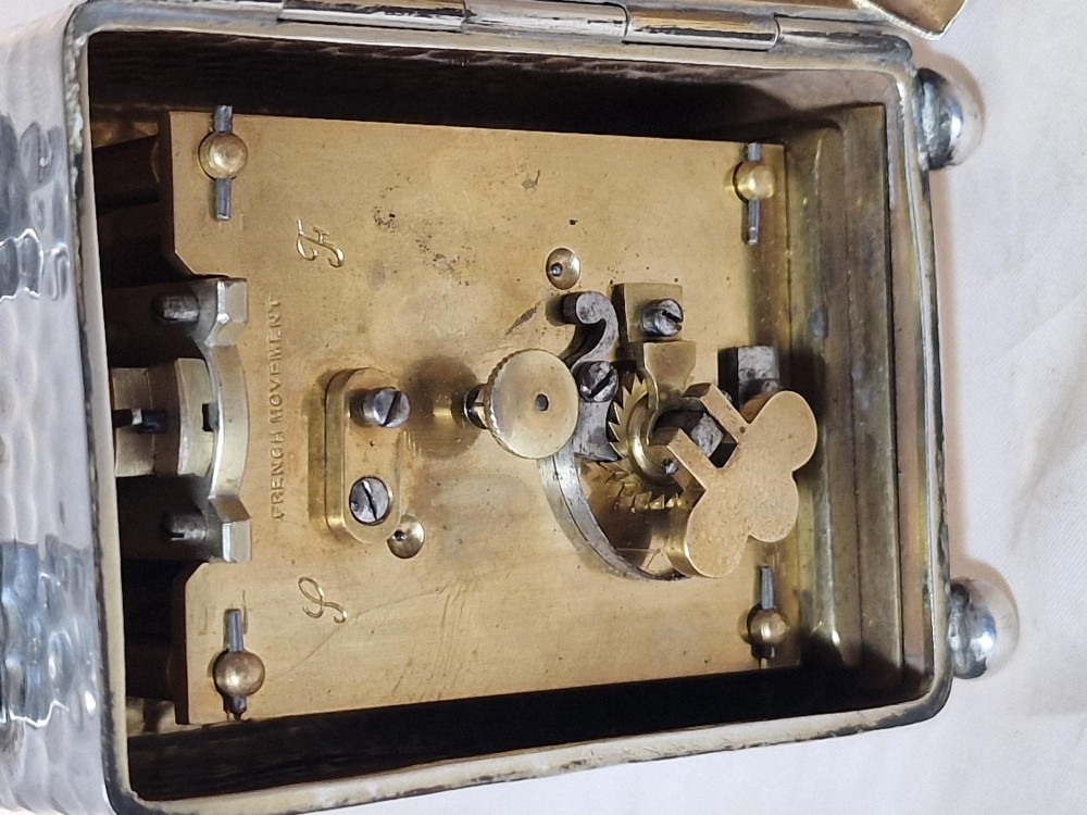 An Edwardian sterling silver carriage clock featuring a planished hammered case, scrolling carrying handle, ball feet, and a white dial with Roman numerals. View V