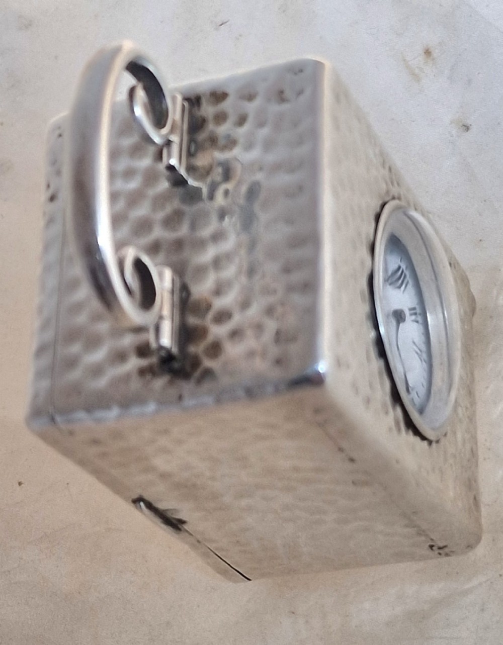 An Edwardian sterling silver carriage clock featuring a planished hammered case, scrolling carrying handle, ball feet, and a white dial with Roman numerals. View VI