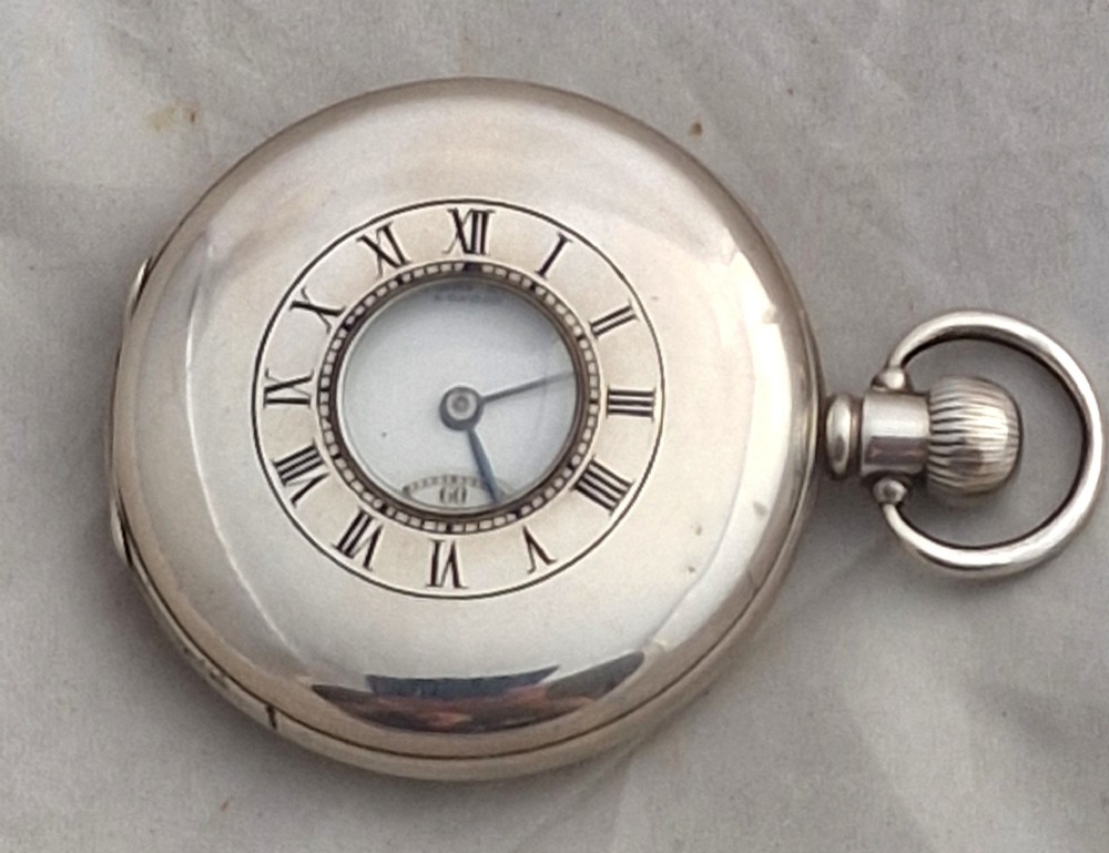 A polished silver half hunter pocket watch displaying an engraved outer ring with black Roman numerals surrounding a central circular glass window.